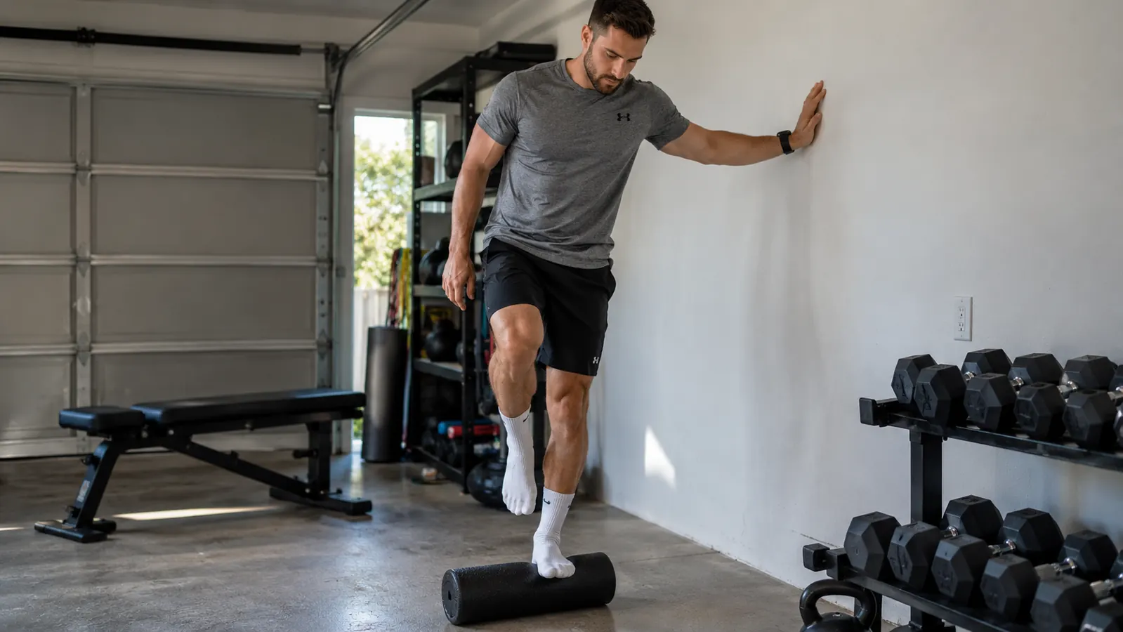 A man in his late fifties stands on a foam roller with one foot, using a wall for balance, in a garage
